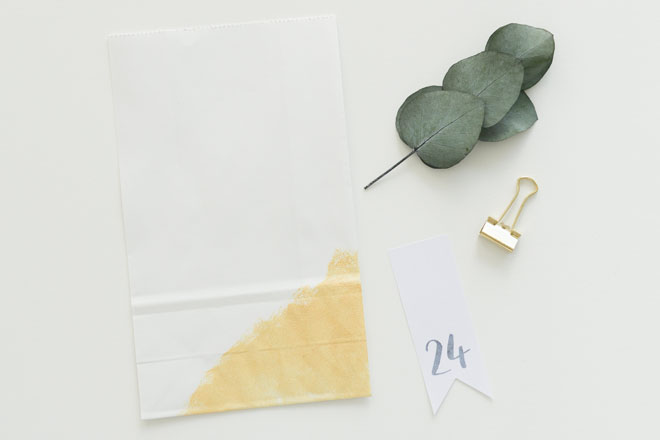 Adventskalender selber machen, vorbereitete weiß-goldene Papiertüte mit Eukalyptuszweig, Zahlenfähnchen und Foldback Klammer