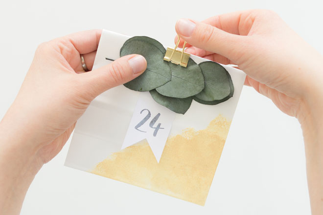 Adventskalender selber machen, Zahlenfähnchen und Eukalyptus an weiß-goldener Papiertüte mit Foldback Klammer befestigen